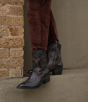 A person wearing Bed|Stü’s Chamise dark, embroidered western boots and brown corduroy pants stands on a concrete ledge next to a brick wall.