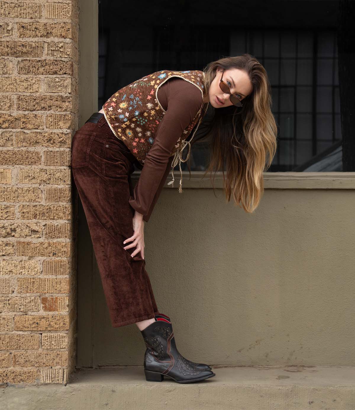 A person wearing sunglasses, a floral vest, brown pants, and stylish Bed|Stü Chamise western boots leans forward against a wall outdoors, looking toward the camera.