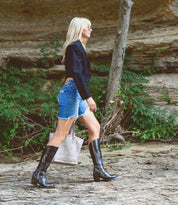 A person with long blond hair, dressed in a black jacket and denim shorts, walks outdoors on rocky ground with greenery in the background, carrying the Bed|Stü Charis beige bag.