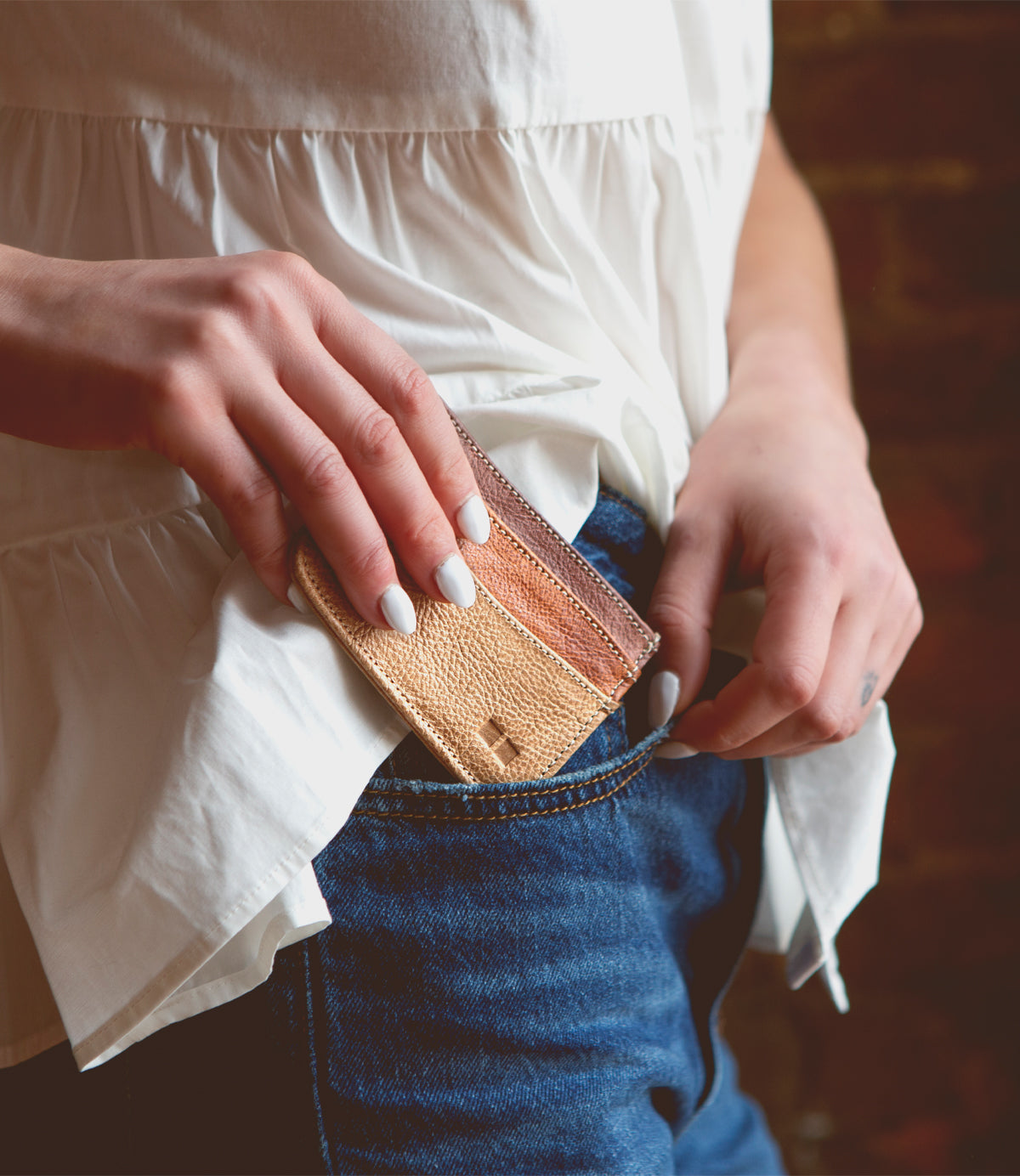 A person with white-painted nails slips the Bed|Stü Chuck beige and brown compact card wallet into the front pocket of blue jeans while wearing a white blouse.