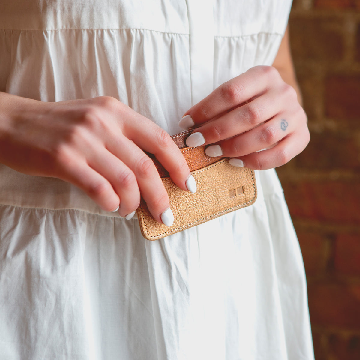 A person with white-painted nails holds the Chuck compact card wallet by Bed|Stü, crafted from tan leather, in both hands. Wearing a white blouse with a blurred brick wall in the background, they showcase this stylish everyday carry essential.