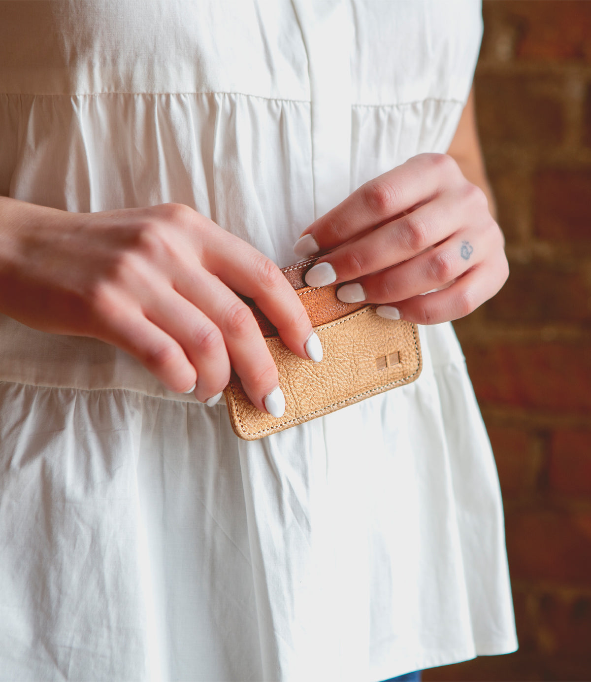 A person with white-painted nails holds the Chuck compact card wallet by Bed|Stü, crafted from tan leather, in both hands. Wearing a white blouse with a blurred brick wall in the background, they showcase this stylish everyday carry essential.