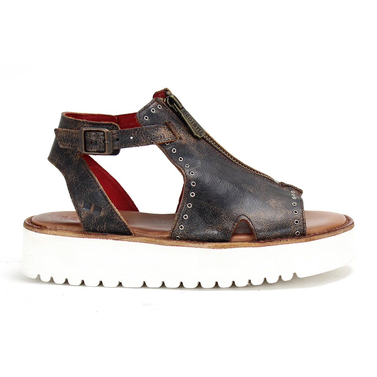 The Bed|Stü Clancy is a single dark brown cutout leather sandal featuring a front zipper, ankle strap with metal buckle, and a thick white ridged sole, shown from the side.