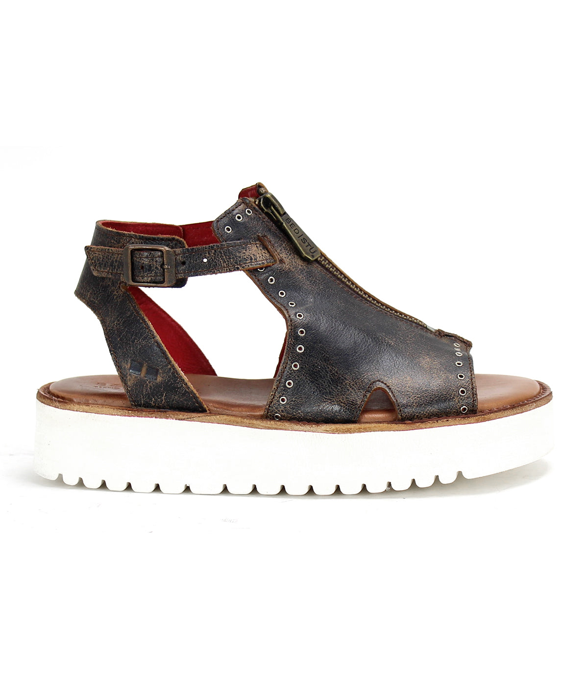 The Bed|Stü Clancy is a single dark brown cutout leather sandal featuring a front zipper, ankle strap with metal buckle, and a thick white ridged sole, shown from the side.