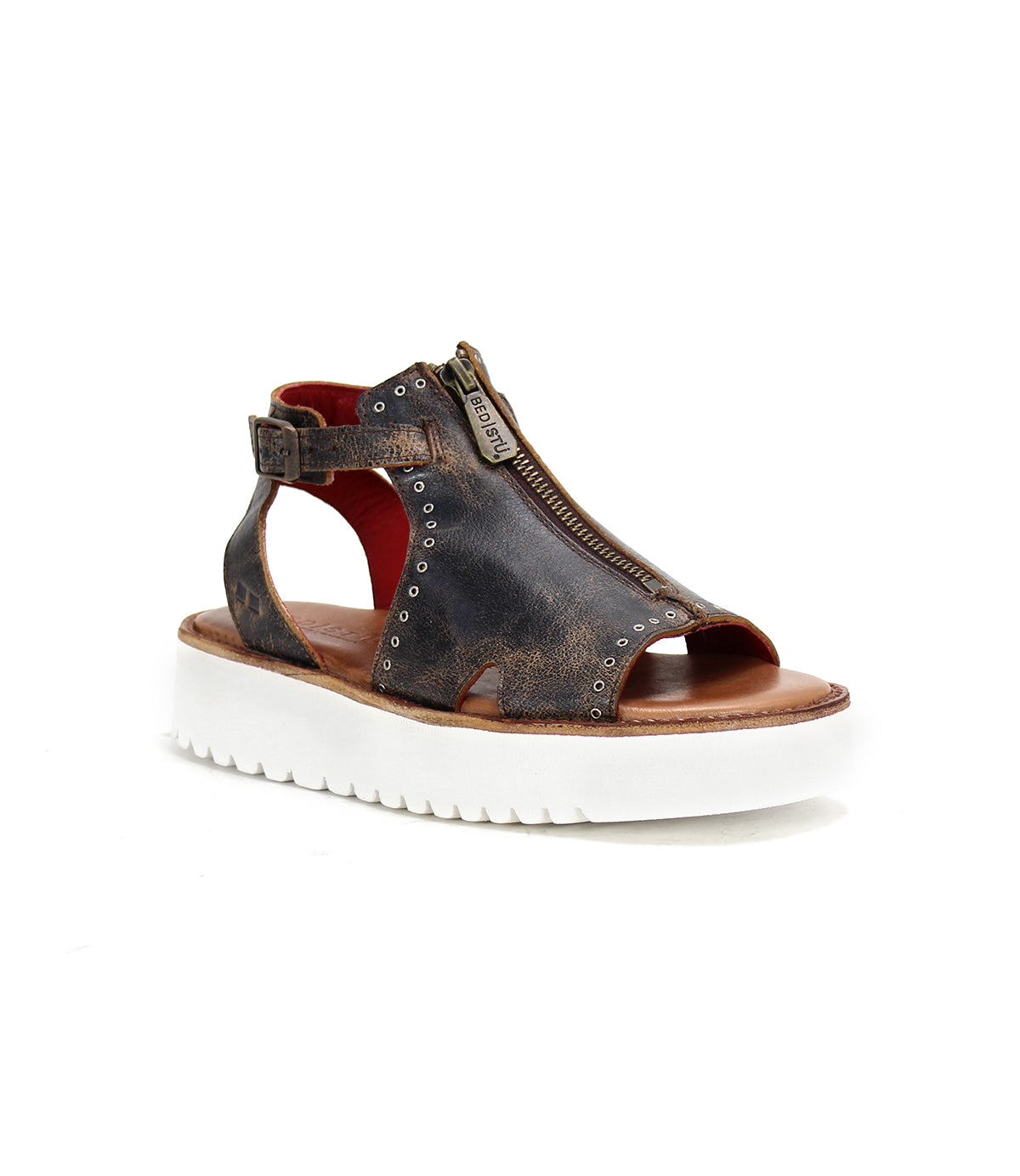 The Bed|Stü Clancy is an open-toe brown sandal, crafted from stylish cutout leather with a front zipper, ankle strap, and metallic rivets. It features a thick white ridged sole and cushioned insole for all-day comfort, shown against a white background.