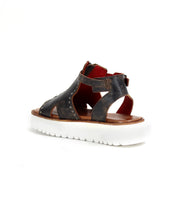 The Bed|Stü Clancy is a black leather platform sandal highlighted by metallic rivet details, cutout leather accents, a vibrant red interior lining, and a convenient back zipper. It features a thick white ridged sole and a cushioned insole for enhanced comfort, presented against a plain white background.