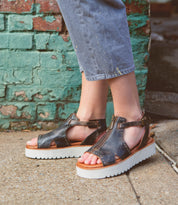 Wearing the Clancy black leather platform sandals with a cushioned insole from Bed|Stü, paired with gray pants, a person stands on a sidewalk near a weathered brick wall.