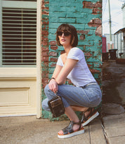 A woman in sunglasses, a white t-shirt, and jeans squats by a weathered brick wall outdoors, holding a small black bag. She completes her look with the Bed|Stü Clancy platform shoes, featuring a cushioned insole for all-day comfort.