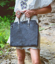 A person in a white outfit stands outdoors holding the Bed|Stü Commuter—a large black leather bag featuring silver hardware and beige handles.