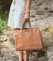 A person in light denim shorts stands outdoors on a rocky surface, holding the Bed|Stü Commuter bag in large tan leather.