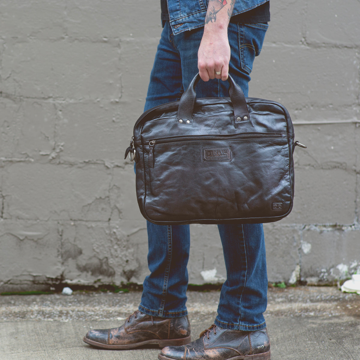 A person dressed in blue jeans, a denim jacket, and brown shoes is holding the Bed|Stü Conclusion— a vegetable-tanned leather messenger bag featuring a dedicated laptop section—while standing on a concrete sidewalk in front of a gray wall.