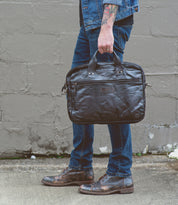 A person dressed in blue jeans, a denim jacket, and brown shoes is holding the Bed|Stü Conclusion— a vegetable-tanned leather messenger bag featuring a dedicated laptop section—while standing on a concrete sidewalk in front of a gray wall.