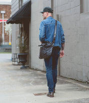 A man in a denim jacket, blue jeans, and cap walks on the sidewalk beside a gray building, carrying the Bed|Stü Conclusion messenger bag crafted from dark vegetable-tanned leather and featuring a convenient laptop section.