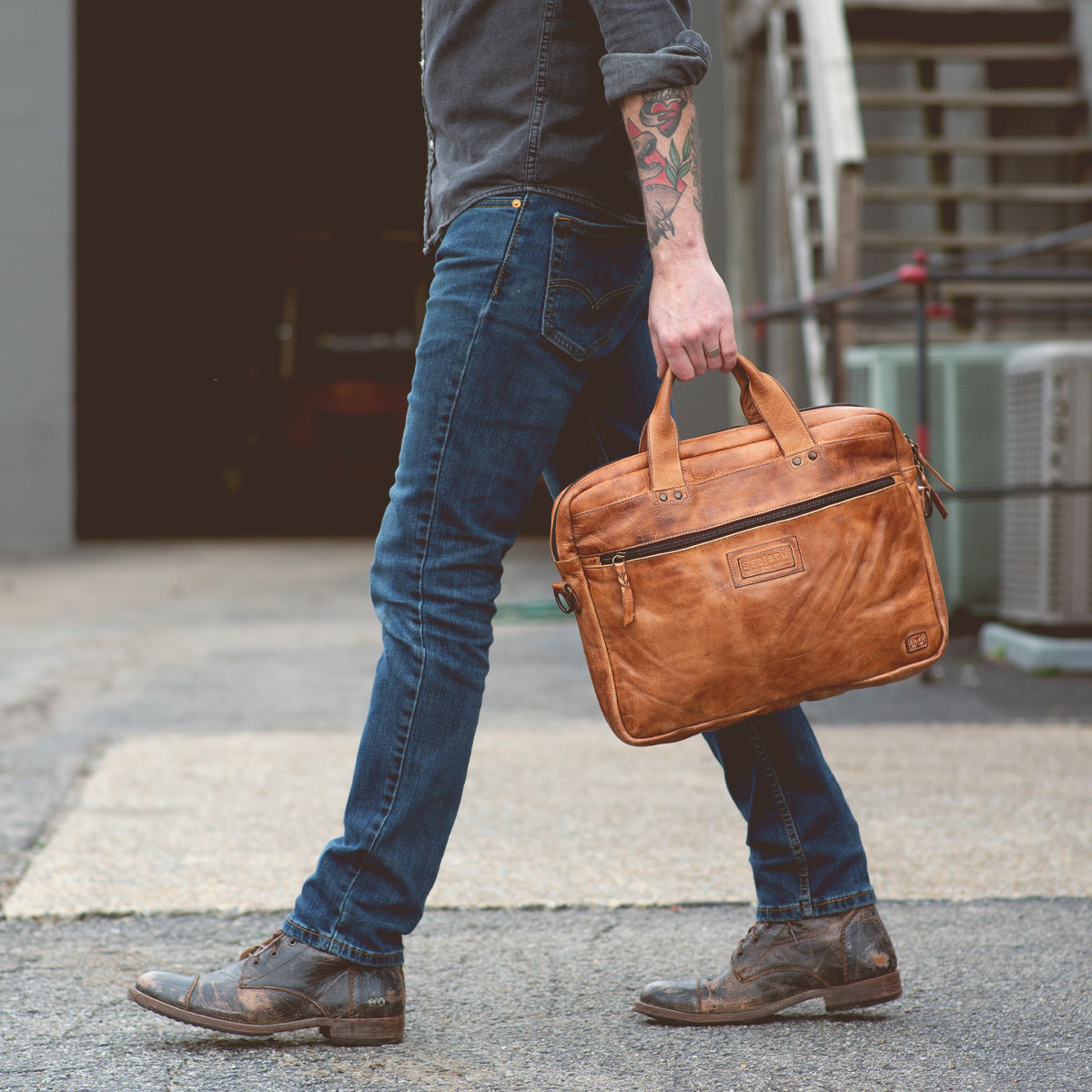 A person wearing jeans and boots walks outside on pavement, holding Bed|Stü’s Conclusion messenger bag, crafted from brown vegetable-tanned leather and featuring a dedicated laptop section. Only the lower half of the body is visible.
