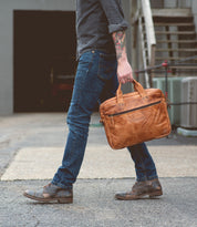 A person wearing jeans and boots walks outside on pavement, holding Bed|Stü’s Conclusion messenger bag, crafted from brown vegetable-tanned leather and featuring a dedicated laptop section. Only the lower half of the body is visible.