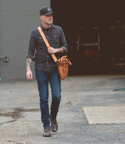A man wearing a baseball cap, dark shirt, jeans, and boots walks outside while carrying the Bed|Stü Conclusion vegetable-tanned leather messenger bag, featuring a spacious laptop section.