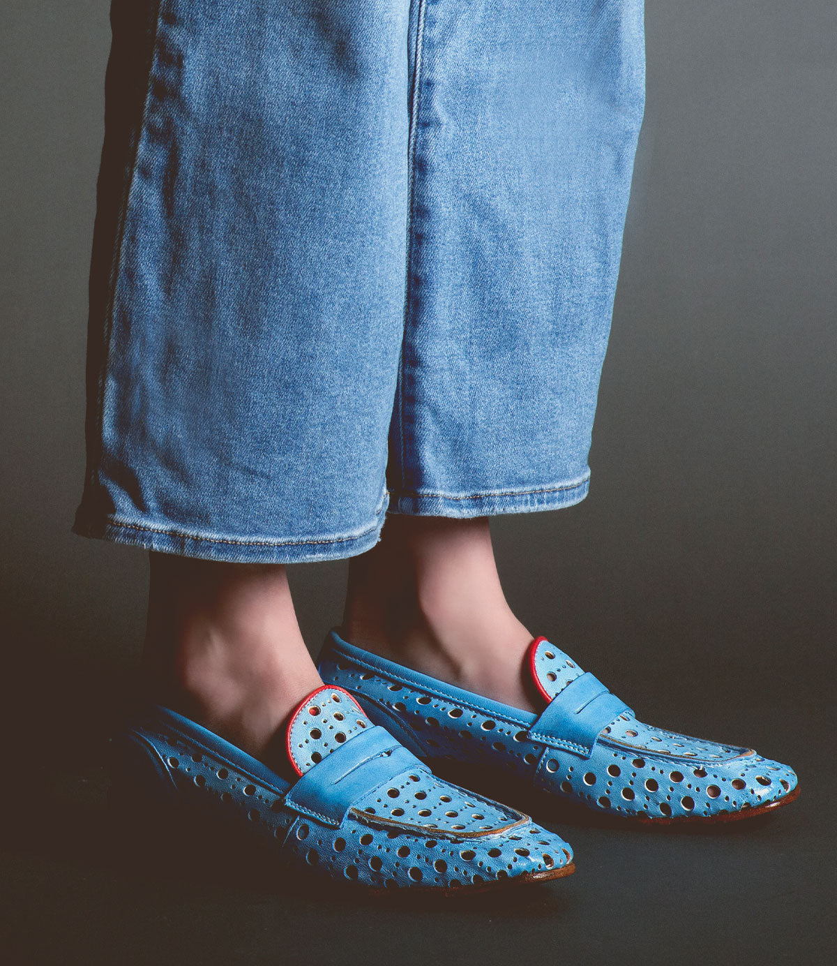 Wearing wide-leg blue jeans and Cornflower—stylish women's loafers by Bed|Stü, handcrafted in Italy in light blue leather with perforations and red accents—the person stands against a dark background.
