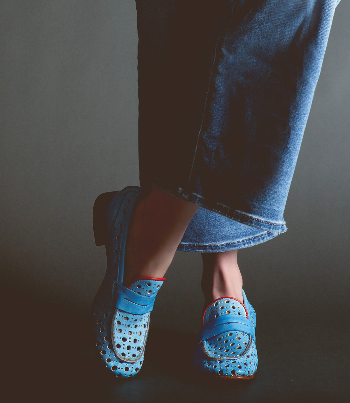 A person wearing the Bed|Stü Cornflower blue perforated women’s loafers, handcrafted in Italy, paired with cropped blue jeans stands against a dark background, one foot slightly raised and crossed in front of the other.
