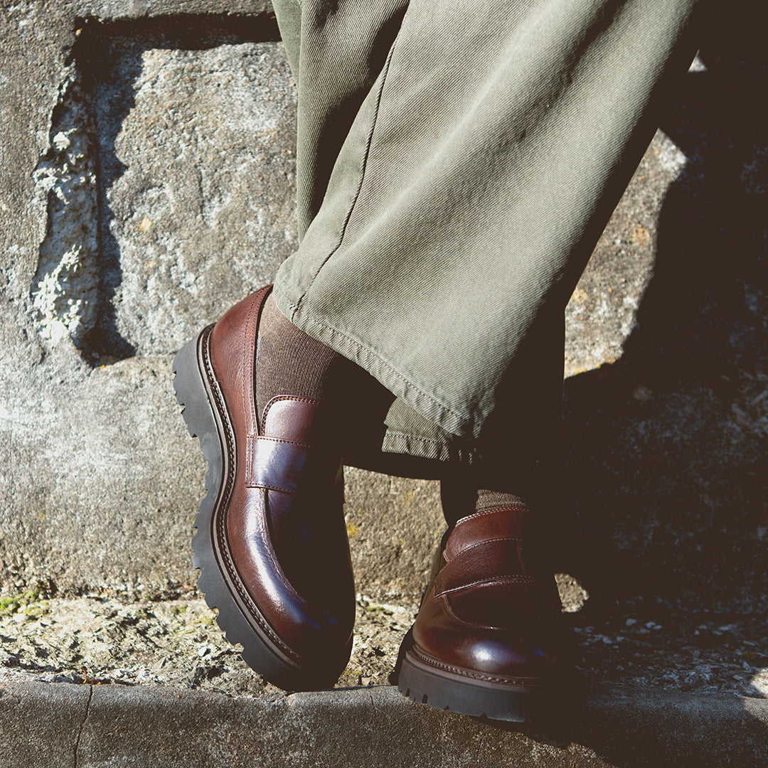 A person stands against a stone wall wearing olive green pants, brown socks, and the Bed|Stü Costruito—Italian-made penny loafers crafted from vegetable-tanned leather and finished with a chunky Livetech outsole.