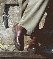 A person stands against a stone wall wearing olive green pants, brown socks, and the Bed|Stü Costruito—Italian-made penny loafers crafted from vegetable-tanned leather and finished with a chunky Livetech outsole.