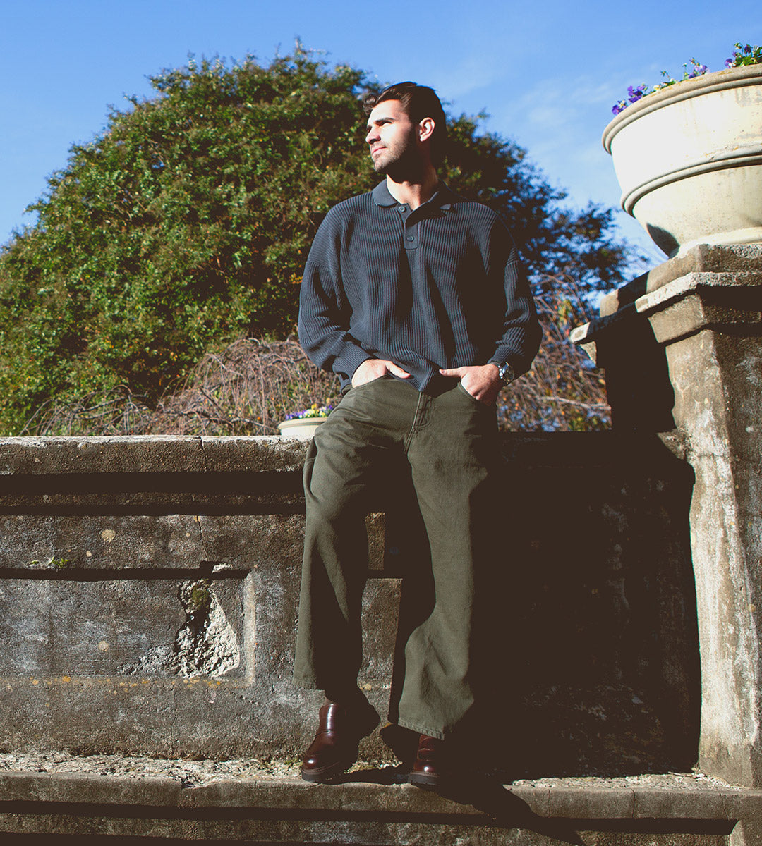 A man stands outdoors against a stone wall, hands in his pockets, dressed in a dark sweater and olive pants, complemented by Bed|Stü’s Costruito Italian-made penny loafers featuring a chunky Livetech outsole, with greenery and a planter visible behind him.