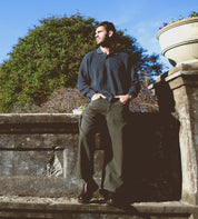 A man stands outdoors against a stone wall, hands in his pockets, dressed in a dark sweater and olive pants, complemented by Bed|Stü’s Costruito Italian-made penny loafers featuring a chunky Livetech outsole, with greenery and a planter visible behind him.