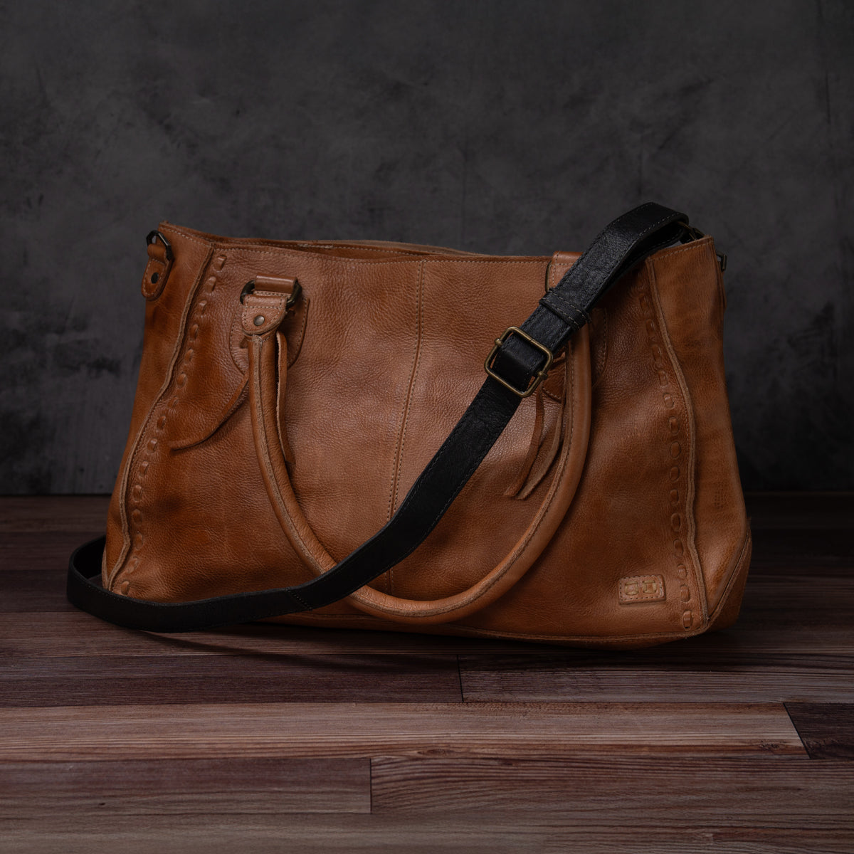 Bed|Stü Crossbody Strap in brown leather, featuring an adjustable strap ideal for crossbody wear, displayed on a wooden floor with a dark textured backdrop.