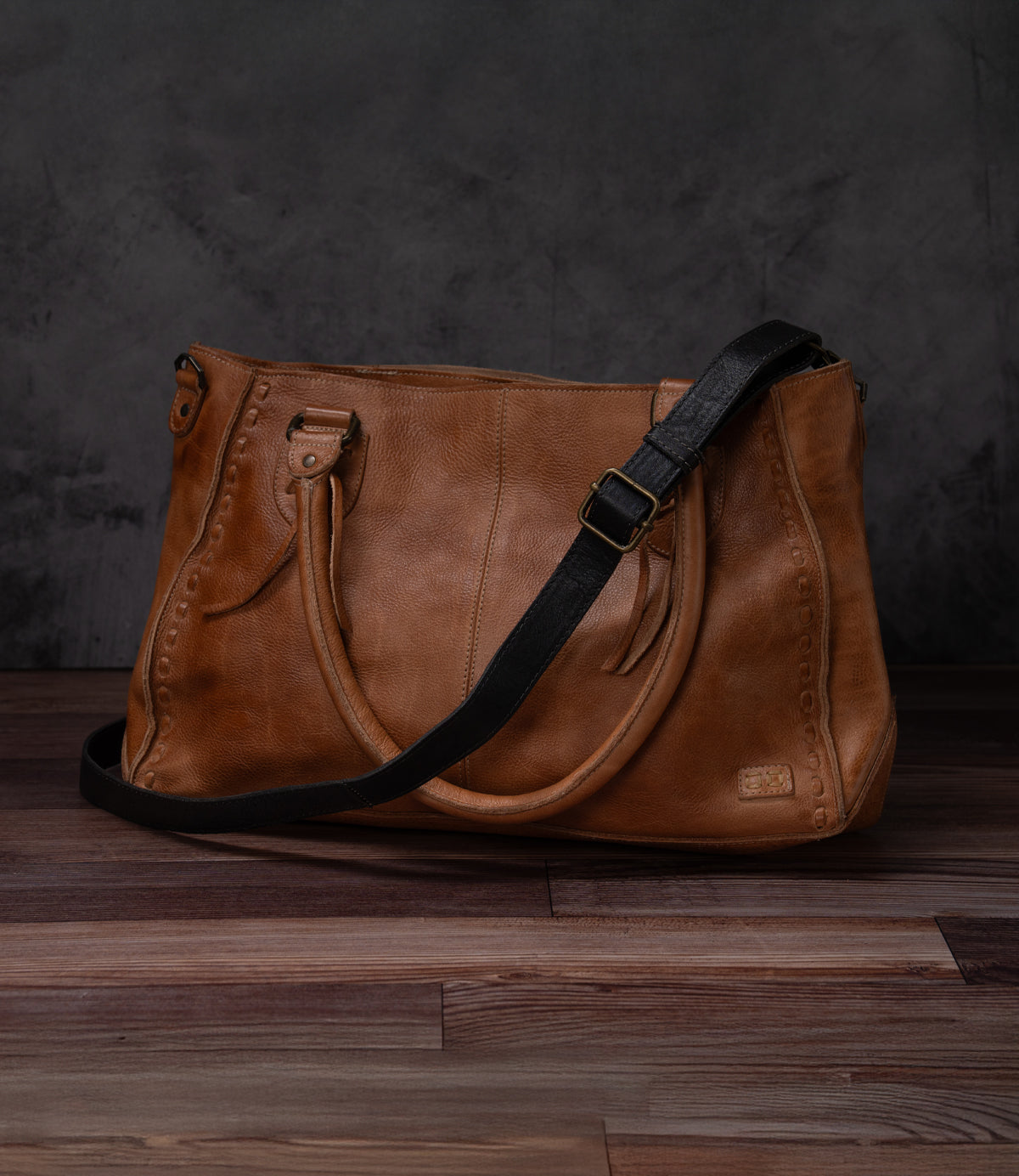 Bed|Stü Crossbody Strap in brown leather, featuring an adjustable strap ideal for crossbody wear, displayed on a wooden floor with a dark textured backdrop.