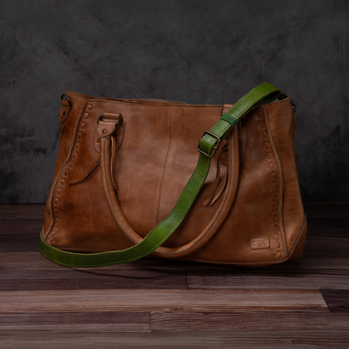 Bed|Stü’s Crossbody Strap features brown leather with dual handles and a green adjustable leather strap, perfect for wearing as a crossbody. Inspired by the timeless charm of Bed|Stü bags, this piece is elegantly displayed on a wooden surface set against a dark gray background.