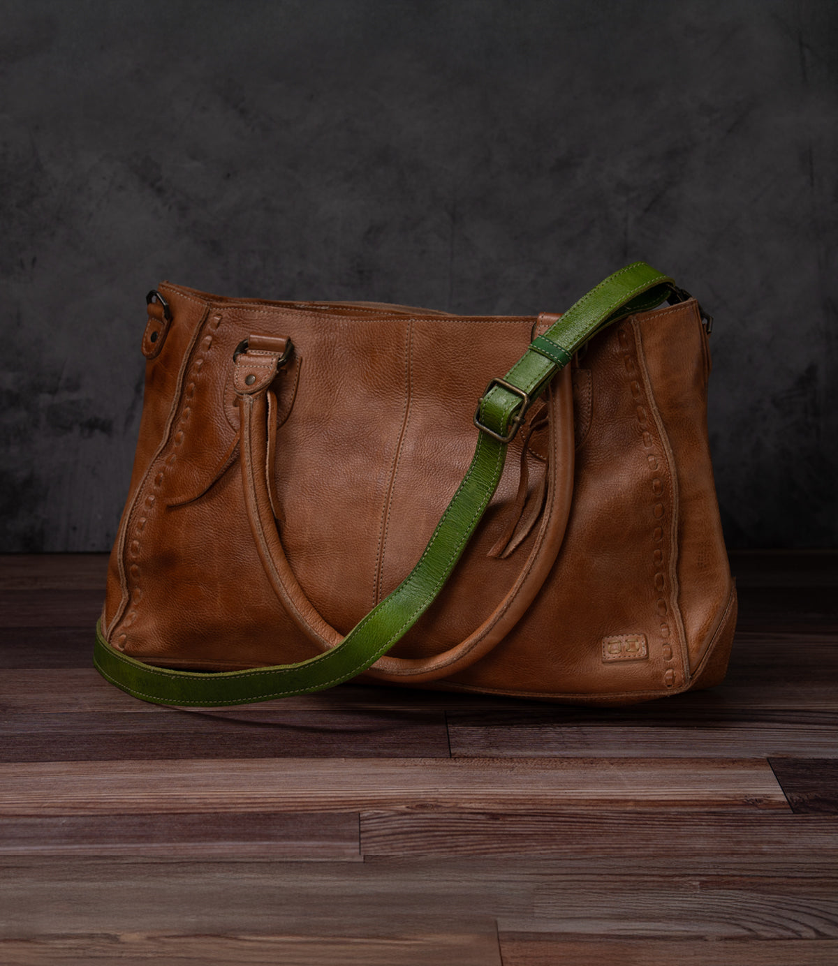 Bed|Stü’s Crossbody Strap features brown leather with dual handles and a green adjustable leather strap, perfect for wearing as a crossbody. Inspired by the timeless charm of Bed|Stü bags, this piece is elegantly displayed on a wooden surface set against a dark gray background.