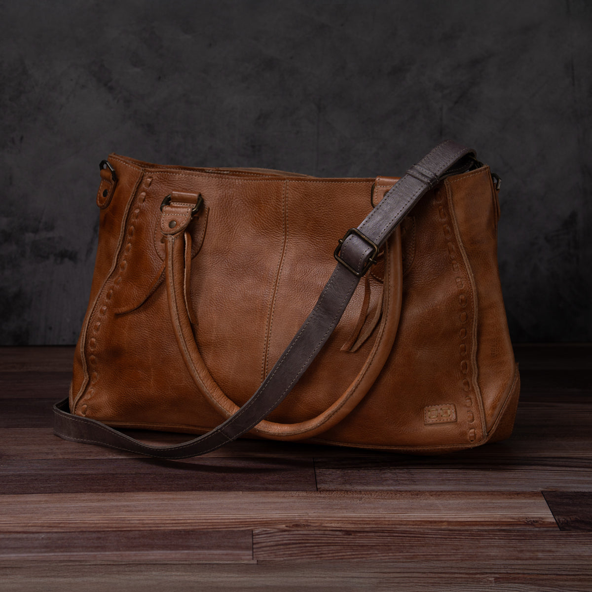 The Bed|Stü Crossbody Strap, crafted from brown leather with double handles and an adjustable leather strap for versatile crossbody wear, is shown on a wooden floor against a dark background—a timeless accessory inspired by classic Bed|Stü designs.