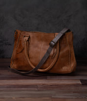 The Bed|Stü Crossbody Strap, crafted from brown leather with double handles and an adjustable leather strap for versatile crossbody wear, is shown on a wooden floor against a dark background—a timeless accessory inspired by classic Bed|Stü designs.