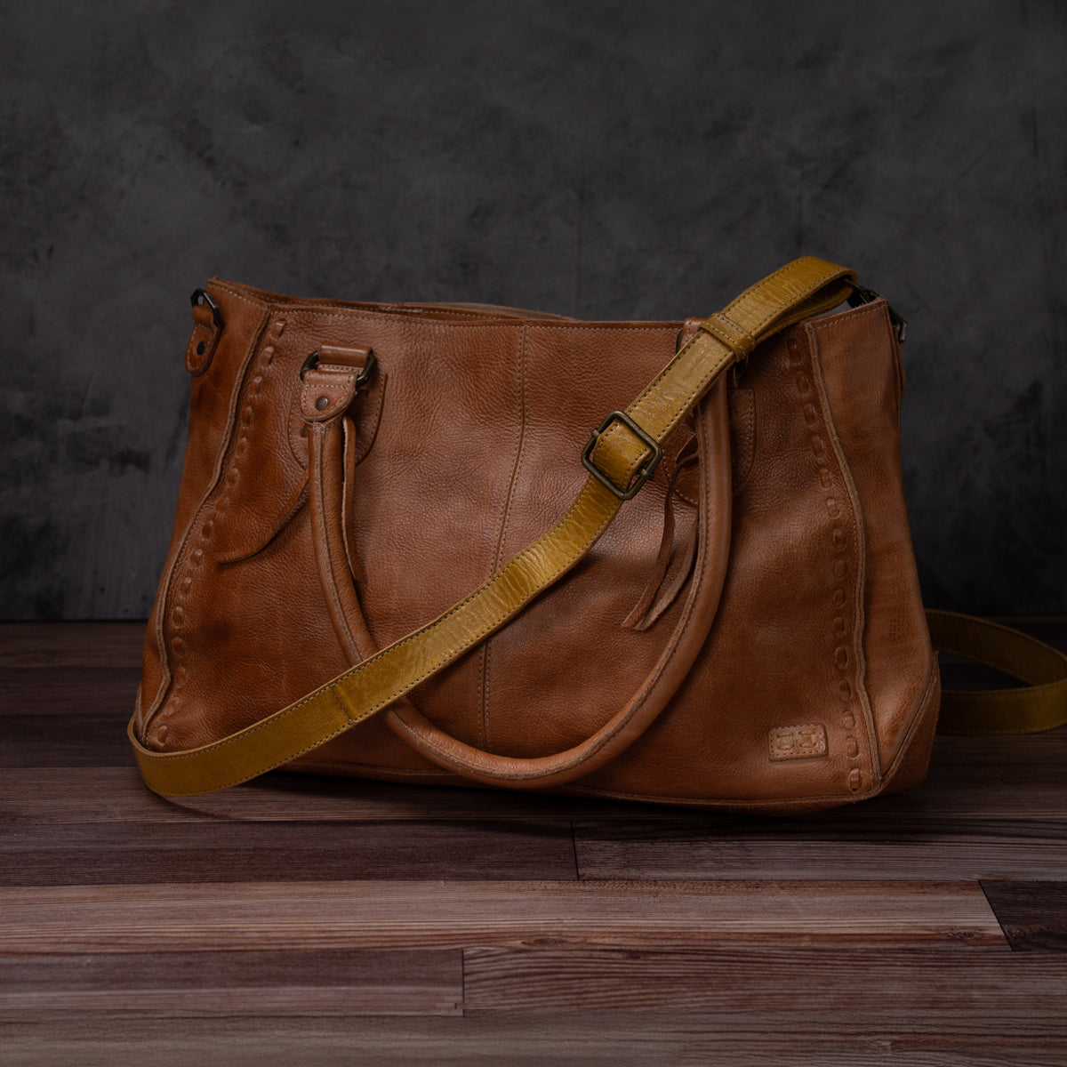 The Bed|Stü Crossbody Strap—a brown leather handbag with a yellow adjustable leather strap—is displayed on a wooden surface against a dark gray textured background.