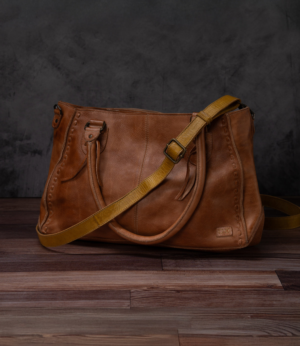 The Bed|Stü Crossbody Strap—a brown leather handbag with a yellow adjustable leather strap—is displayed on a wooden surface against a dark gray textured background.