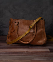 The Bed|Stü Crossbody Strap—a brown leather handbag with a yellow adjustable leather strap—is displayed on a wooden surface against a dark gray textured background.