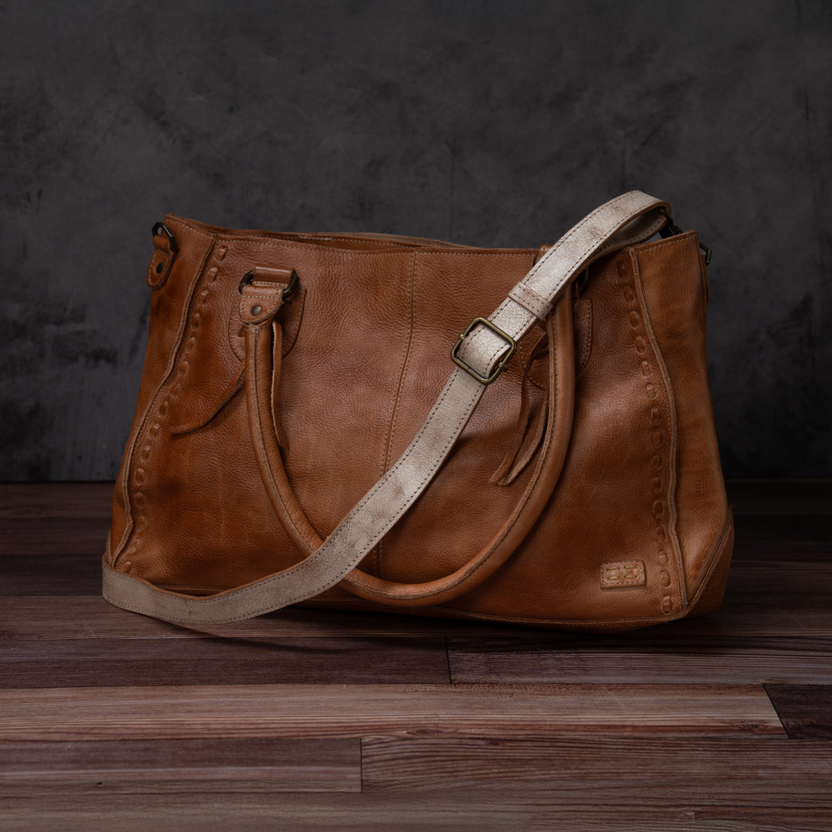 A Bed|Stü Crossbody Strap in brown leather, featuring two handles and an adjustable leather strap for versatile crossbody wear, is displayed on a wooden floor against a dark, textured background, capturing the timeless style of Bed|Stü.