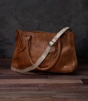 A Bed|Stü Crossbody Strap in brown leather, featuring two handles and an adjustable leather strap for versatile crossbody wear, is displayed on a wooden floor against a dark, textured background, capturing the timeless style of Bed|Stü.