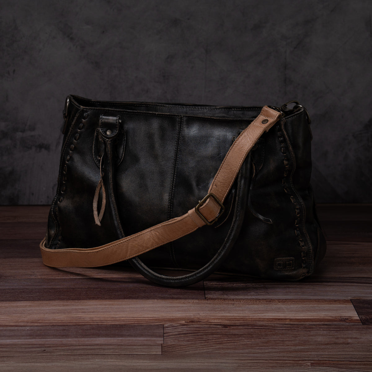 A black leather Bed|Stü Crossbody Strap bag with tan shoulder strap and handles sits on a wooden floor against a dark, textured background, featuring an adjustable leather strap for versatile crossbody wear.