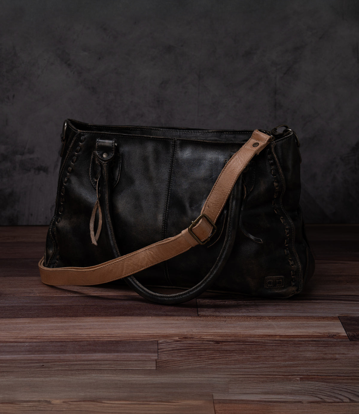 A black leather Bed|Stü Crossbody Strap bag with tan shoulder strap and handles sits on a wooden floor against a dark, textured background, featuring an adjustable leather strap for versatile crossbody wear.