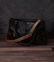 A black leather Bed|Stü Crossbody Strap bag with tan shoulder strap and handles sits on a wooden floor against a dark, textured background, featuring an adjustable leather strap for versatile crossbody wear.