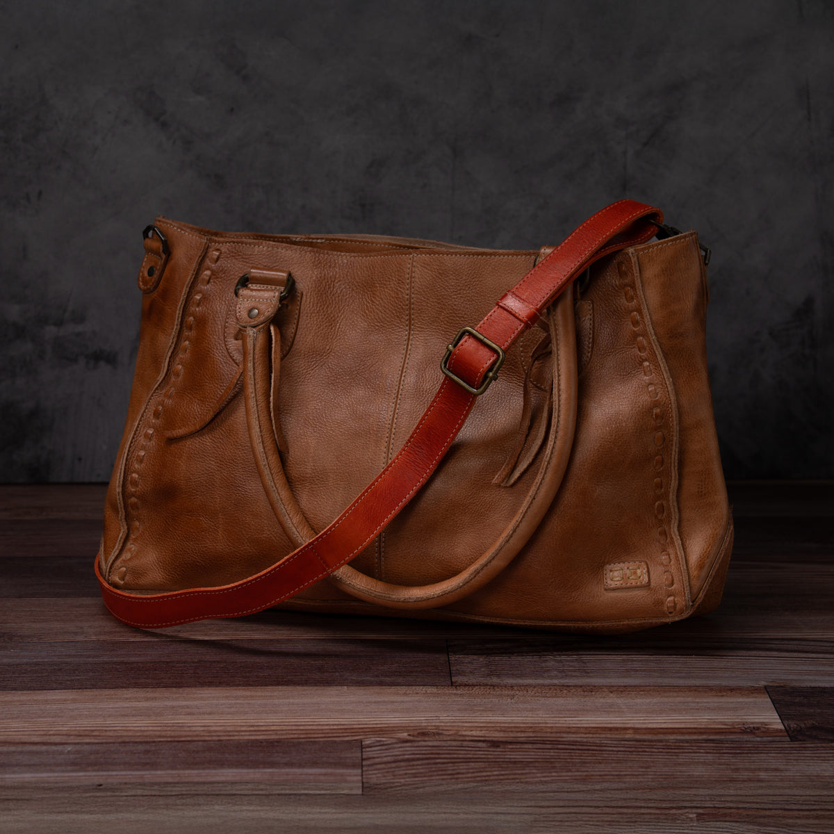 The Bed|Stü Crossbody Strap is shown here attached to a brown leather handbag with double handles and a striking bold red adjustable leather strap for versatile crossbody styling, set on a wooden floor against a dark grey background—perfectly capturing the unique character of Bed|Stü bags.