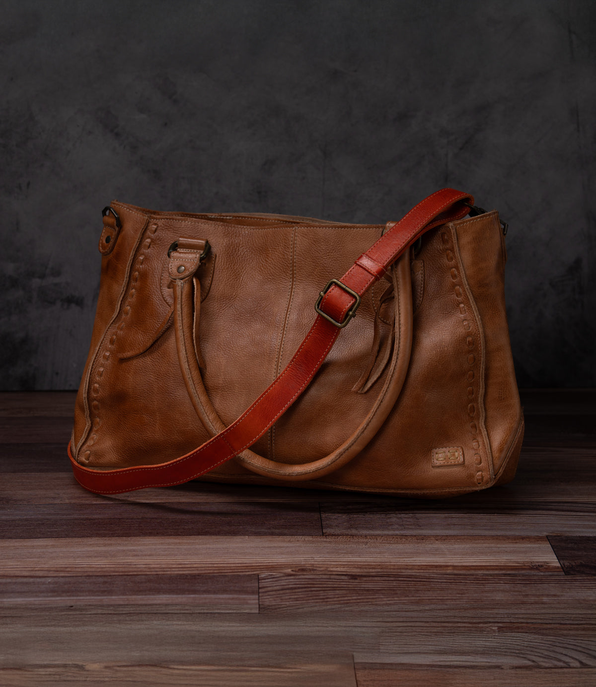 The Bed|Stü Crossbody Strap is shown here attached to a brown leather handbag with double handles and a striking bold red adjustable leather strap for versatile crossbody styling, set on a wooden floor against a dark grey background—perfectly capturing the unique character of Bed|Stü bags.