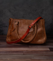 The Bed|Stü Crossbody Strap is shown here attached to a brown leather handbag with double handles and a striking bold red adjustable leather strap for versatile crossbody styling, set on a wooden floor against a dark grey background—perfectly capturing the unique character of Bed|Stü bags.