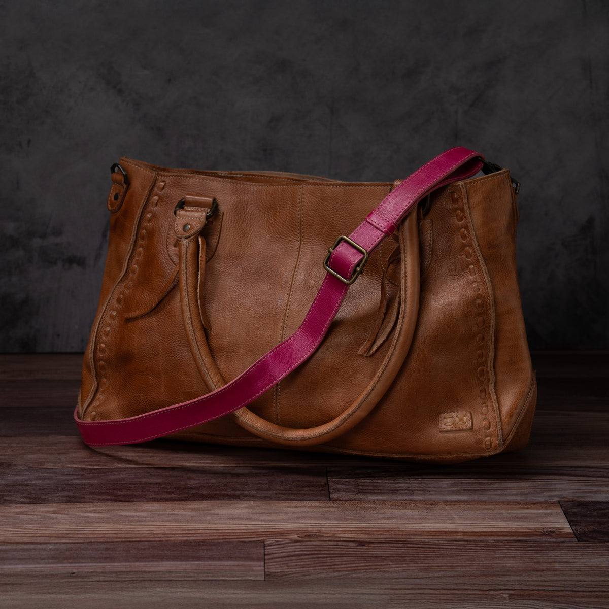 A Bed|Stü Crossbody Strap, crafted from brown leather and featuring two handles and an adjustable leather strap, rests on a wooden floor against a dark background, highlighting the distinctive style of Bed|Stü accessories.
