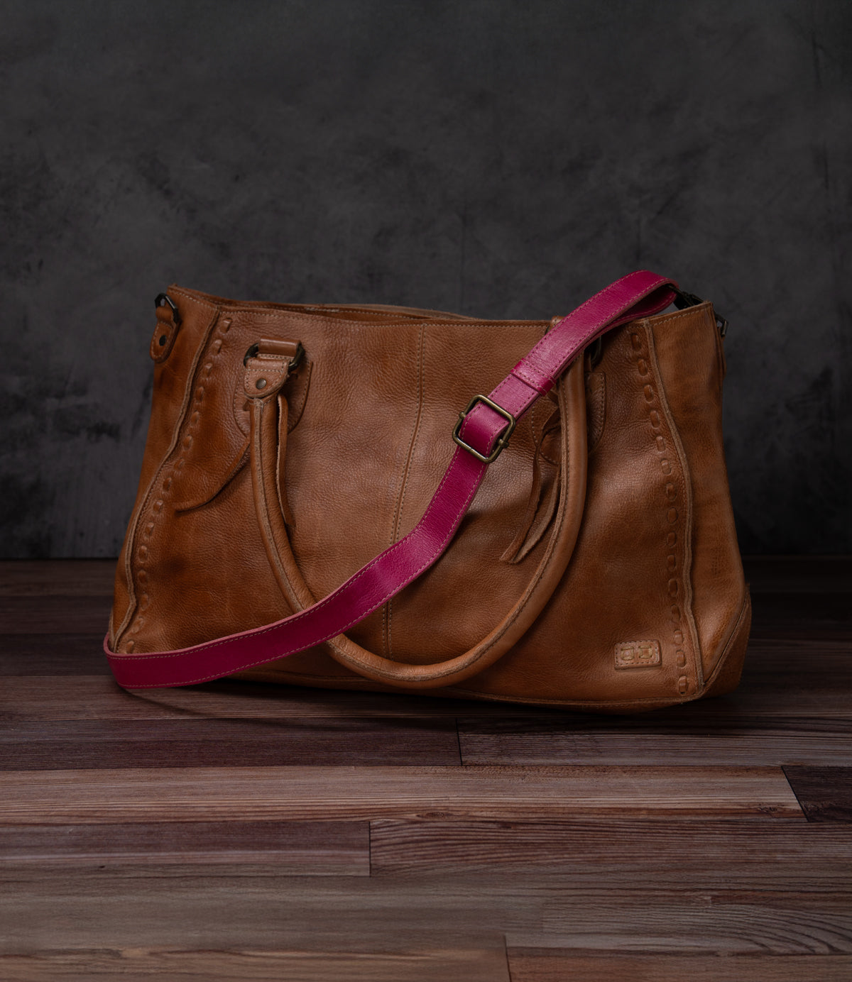 A Bed|Stü Crossbody Strap, crafted from brown leather and featuring two handles and an adjustable leather strap, rests on a wooden floor against a dark background, highlighting the distinctive style of Bed|Stü accessories.