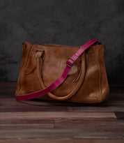 A Bed|Stü Crossbody Strap, crafted from brown leather and featuring two handles and an adjustable leather strap, rests on a wooden floor against a dark background, highlighting the distinctive style of Bed|Stü accessories.