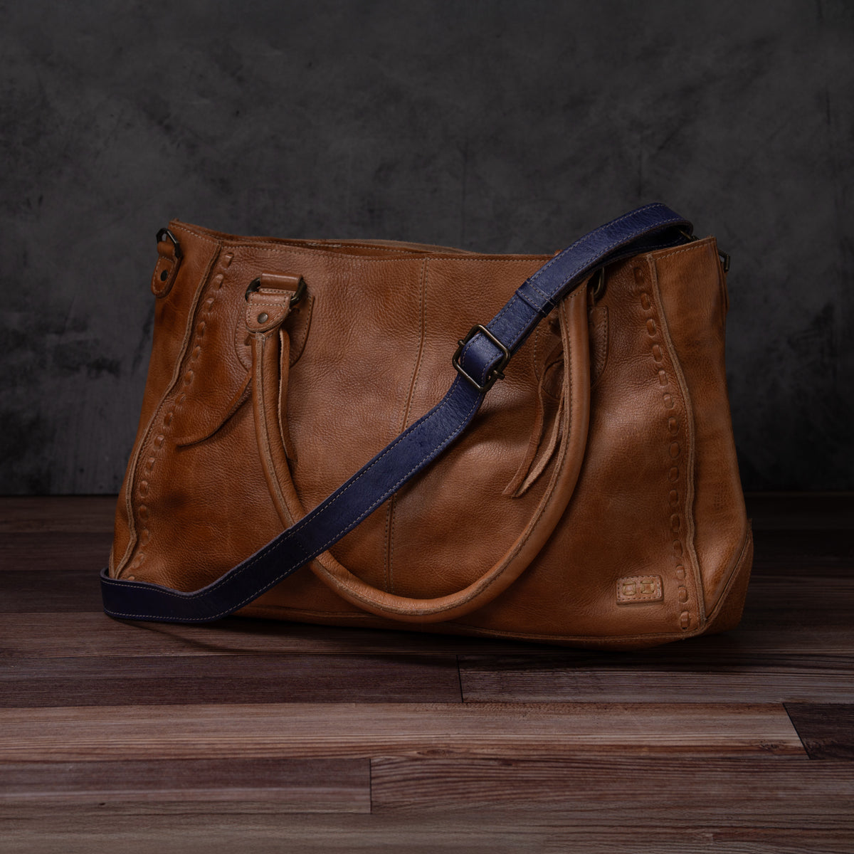 A brown leather Crossbody Strap by Bed|Stü, featuring double handles and a navy blue adjustable leather strap for crossbody wear, is displayed on a wooden floor against a dark textured background. This timeless piece reflects the artisan craftsmanship characteristic of Bed|Stü products.