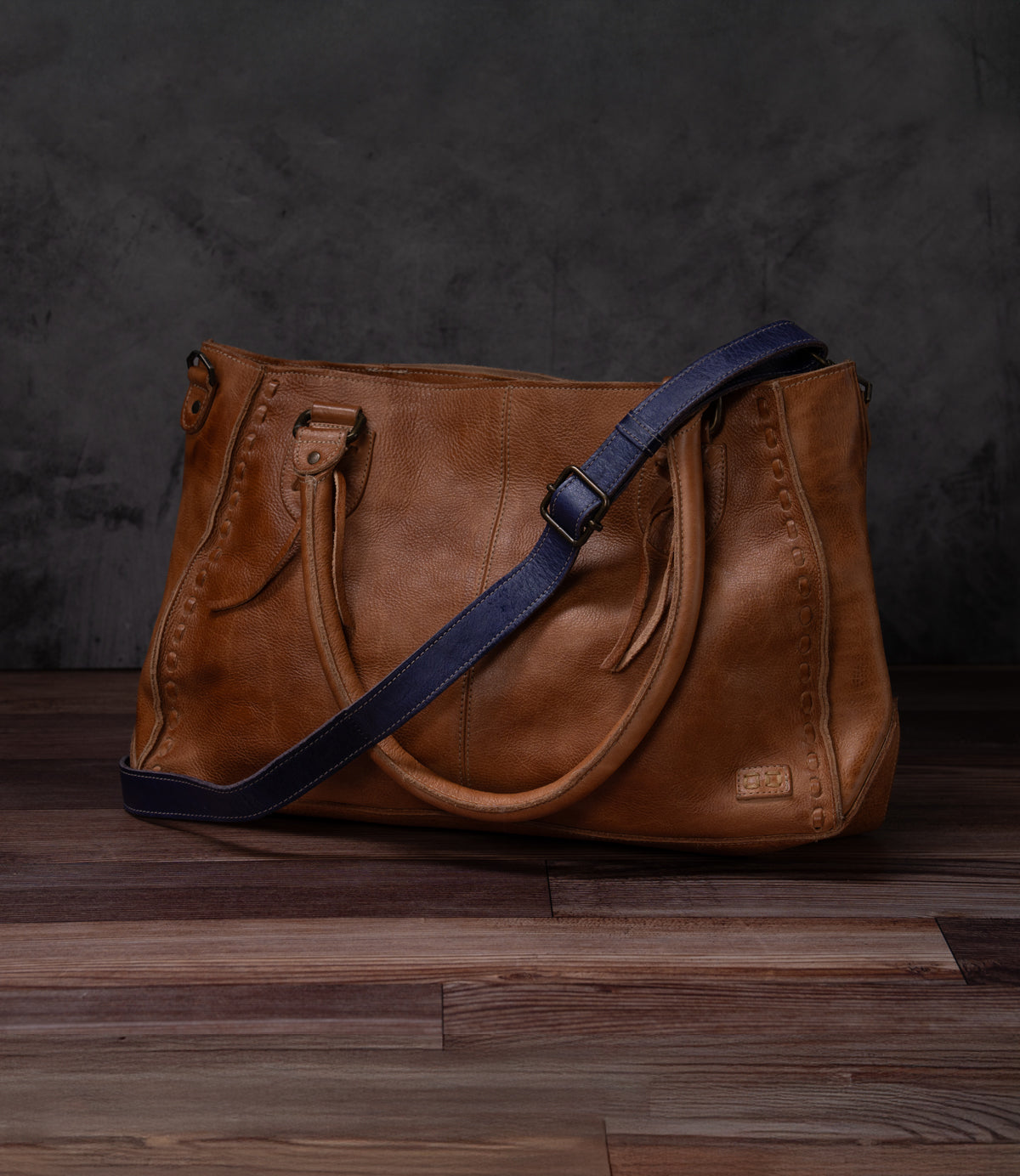 A brown leather Crossbody Strap by Bed|Stü, featuring double handles and a navy blue adjustable leather strap for crossbody wear, is displayed on a wooden floor against a dark textured background. This timeless piece reflects the artisan craftsmanship characteristic of Bed|Stü products.