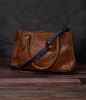A brown leather Crossbody Strap by Bed|Stü, featuring double handles and a navy blue adjustable leather strap for crossbody wear, is displayed on a wooden floor against a dark textured background. This timeless piece reflects the artisan craftsmanship characteristic of Bed|Stü products.