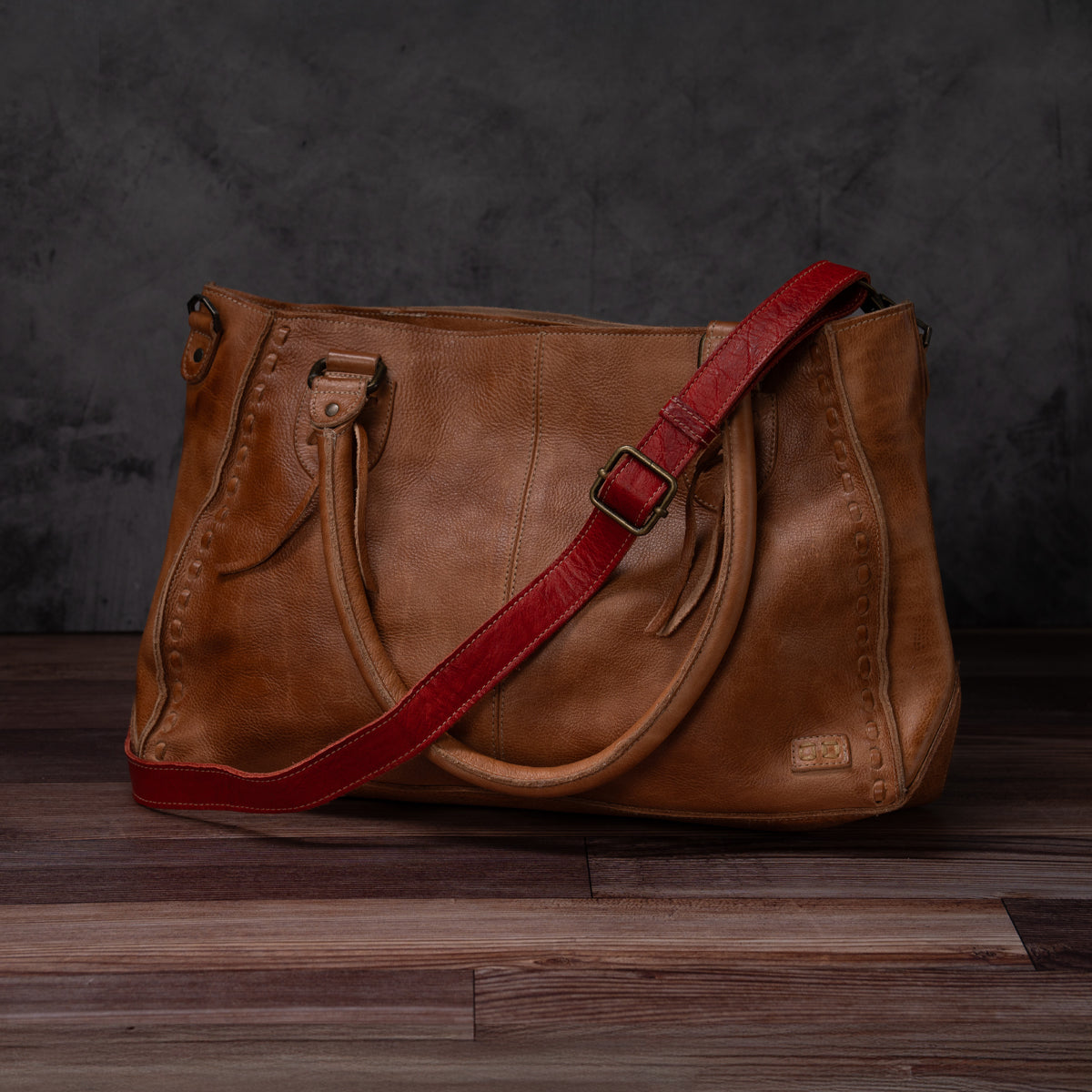 A timeless Bed|Stü Crossbody Strap in brown leather with decorative stitching, two handles, and a detachable red adjustable leather strap for crossbody wear—styled on a wooden floor against a dark textured background.
